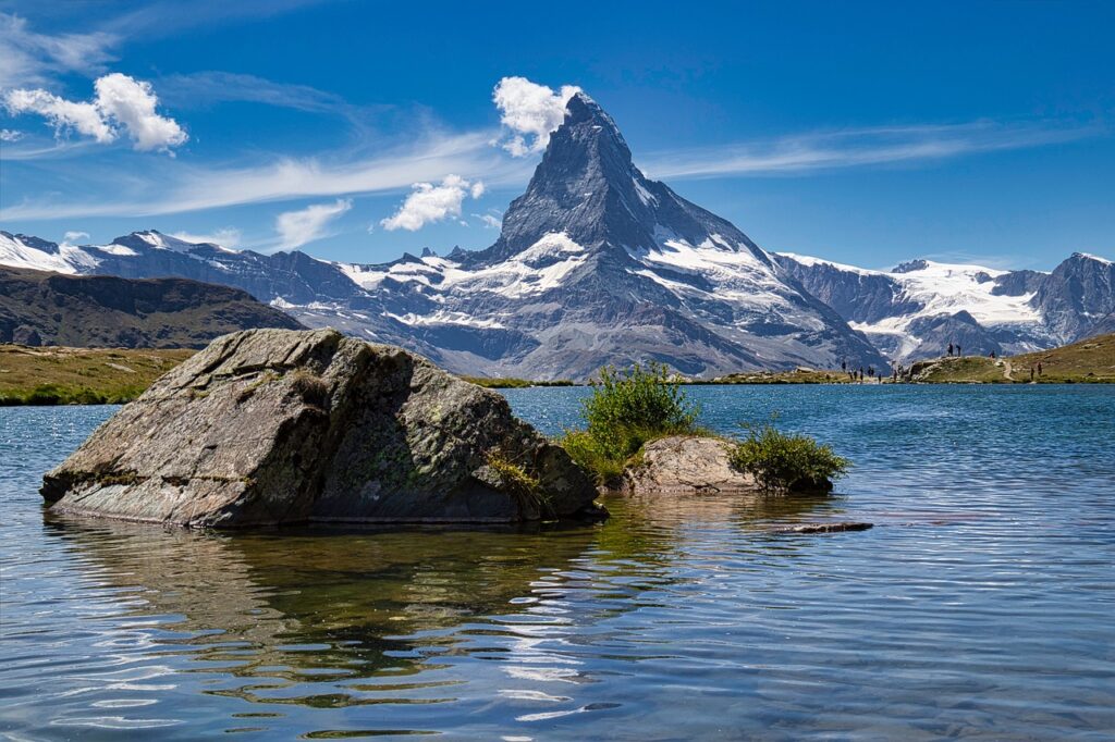 View of the Matterhorn mountain towering over a serene lake, with clear blue skies, surrounding snow-capped peaks, and hikers in the distance