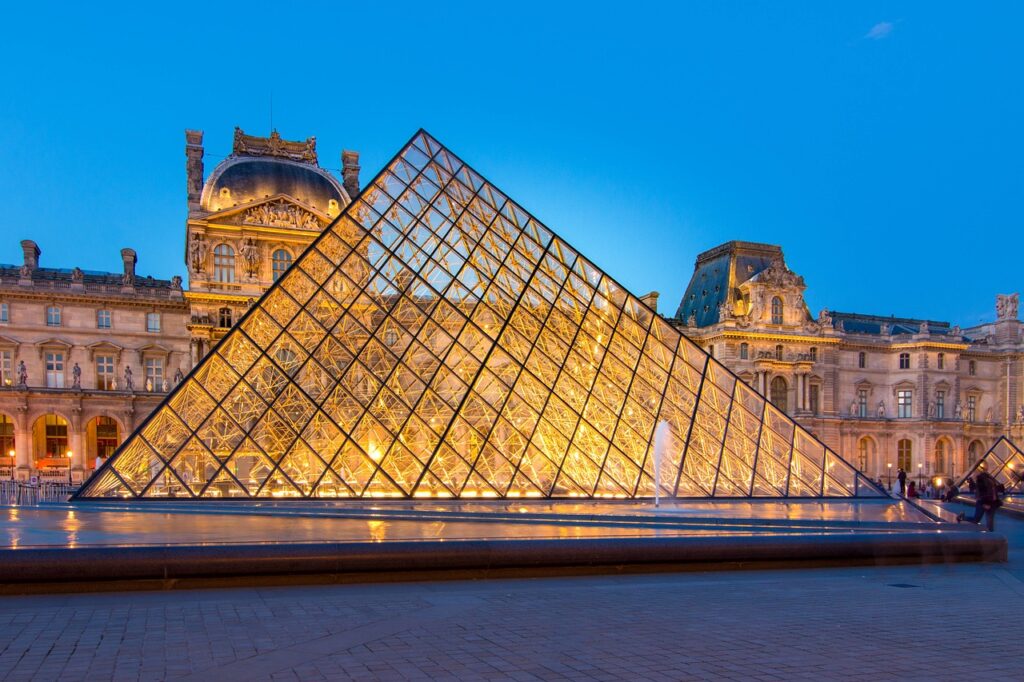 The iconic glass pyramid at the Louvre Museum in Paris illuminated at night, with the historic museum building in the background
