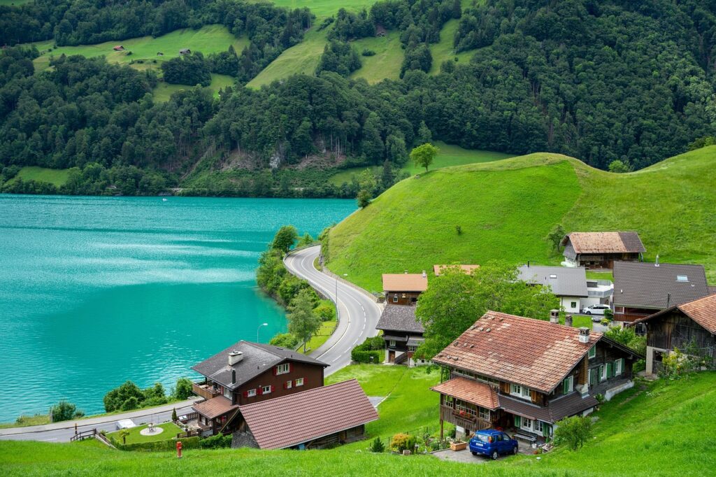 Scenic view of a Swiss village with traditional houses along a road by a turquoise lake, surrounded by lush green hills and dense forest