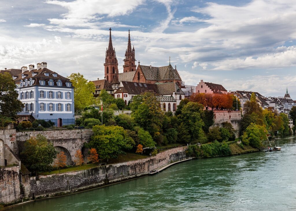 View of Basel’s Old Town with the Munster Cathedral towering over the Rhine River, surrounded by lush greenery and traditional Swiss architecture.